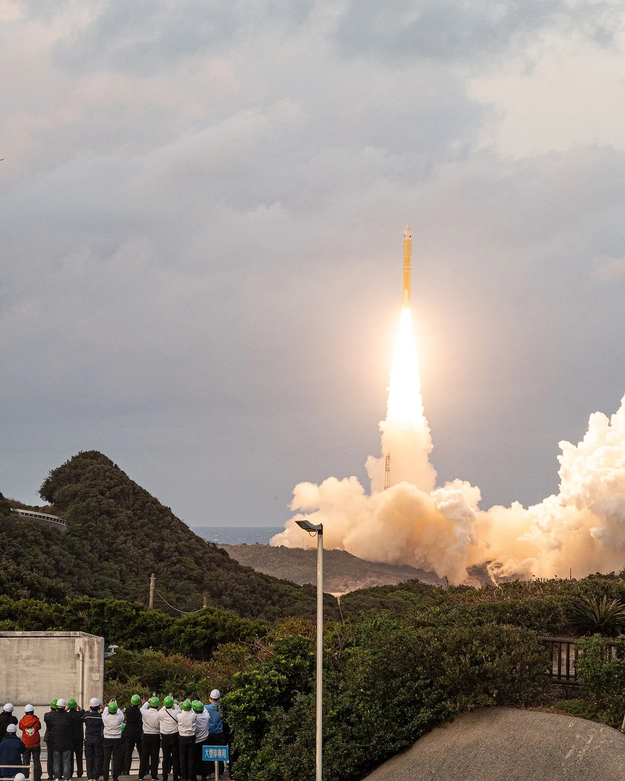 A rocket climbs on a bright column of flame above a green hillside as a group of people in hard hats watch from a viewing area
