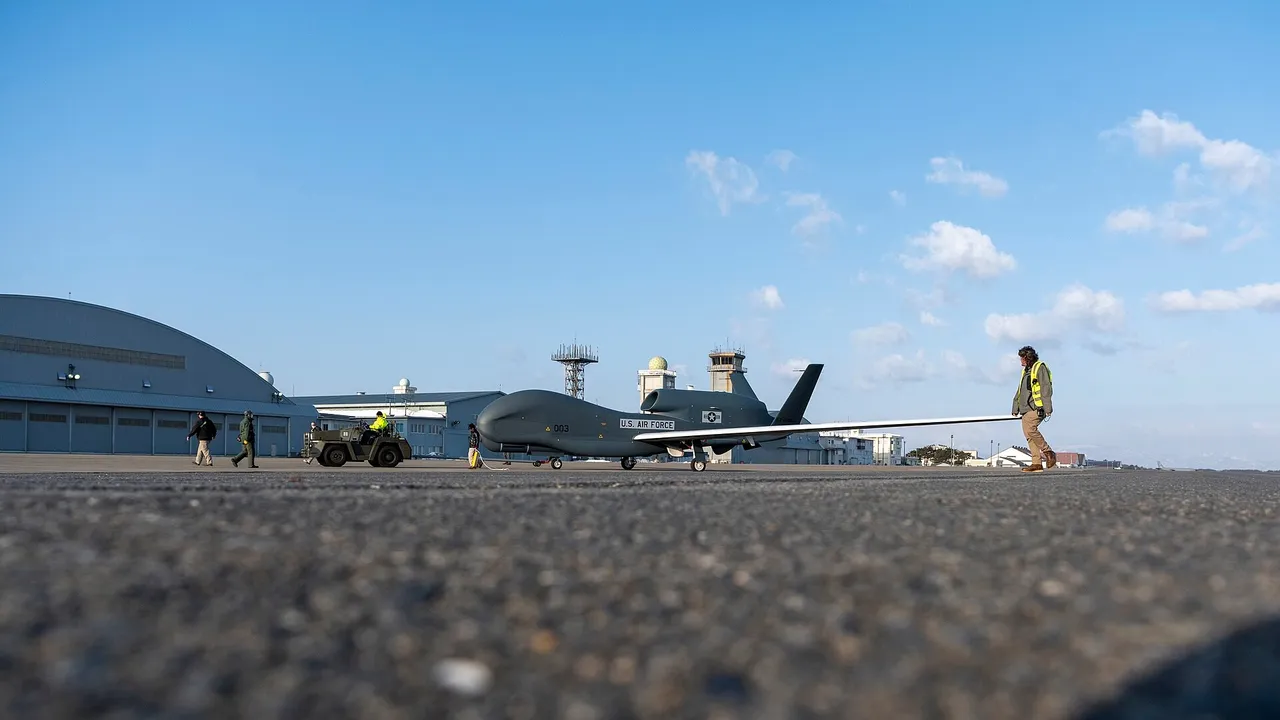 A grey long-winged RQ-4B Global Hawk drone is towed across an air base apron past hangars as ground crew walk alongside