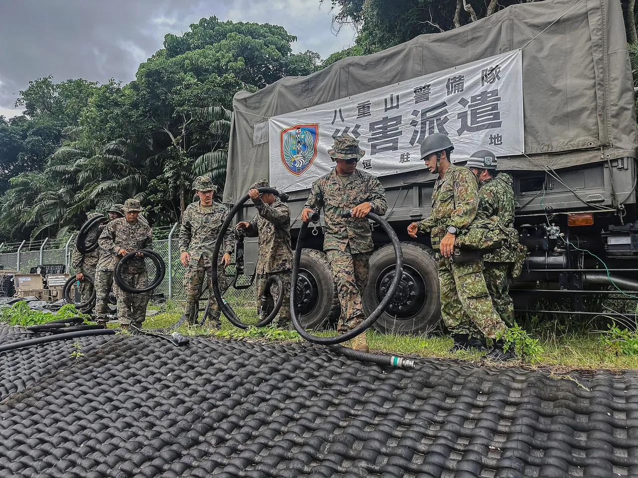 Soldiers in camouflage uniforms carry long water intake hoses across a grassy area at a military camp