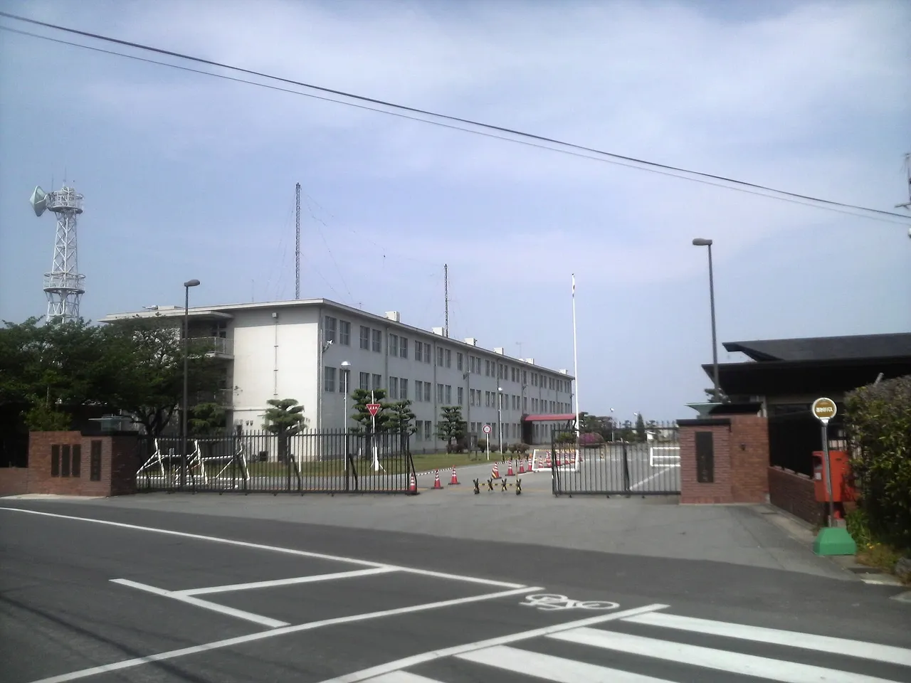 The fenced main gate of a Japanese army camp with a brick gatepost, a three-storey white headquarters building and a radio mast behind it