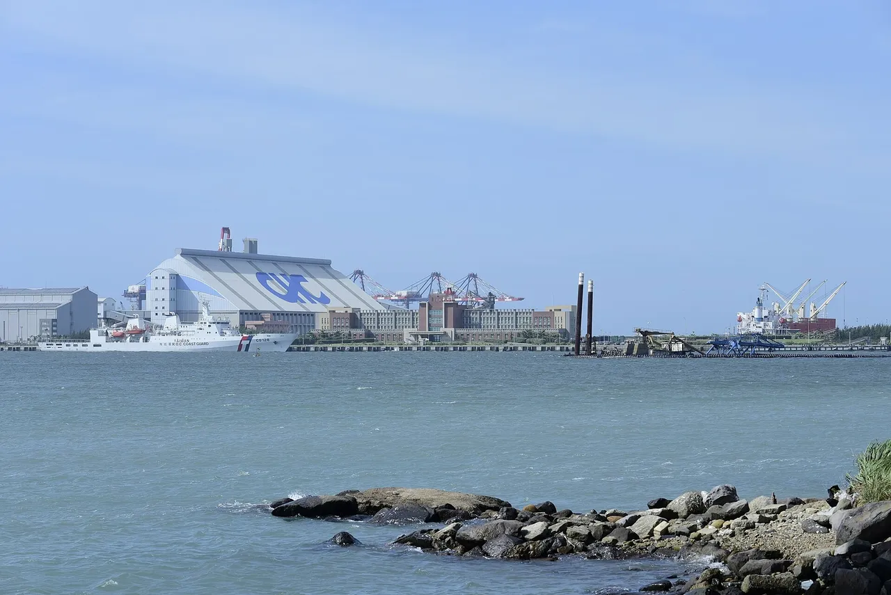 A white Taiwan coast guard ship moored in front of a large warehouse and container cranes across choppy blue-green water, with rocks in the foreground