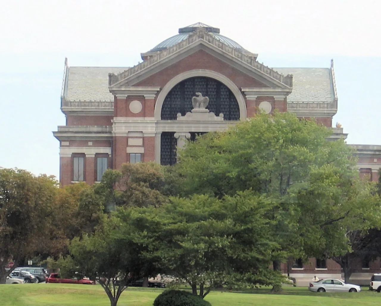 A red-brick neoclassical building with a domed roof and a large arched window topped by an eagle sculpture, partly hidden behind green trees