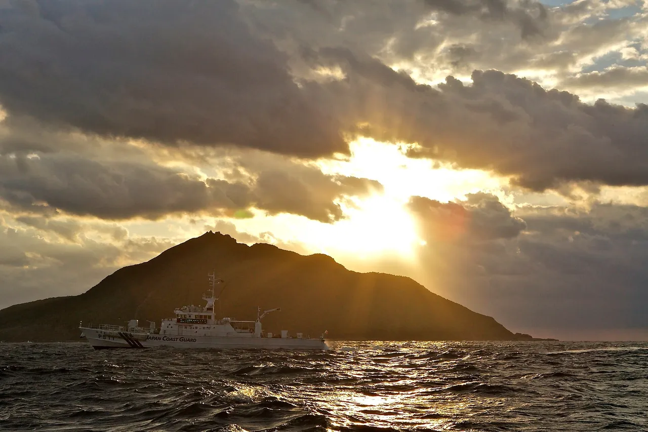 A white Japan Coast Guard patrol ship sails past a dark mountainous island under a cloudy sky with the sun breaking through