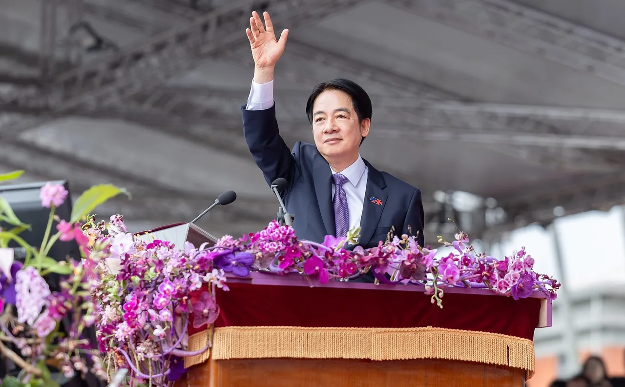 A man in a dark suit and purple tie waves with his right hand raised from a podium decorated with purple and pink orchids under a large canopy