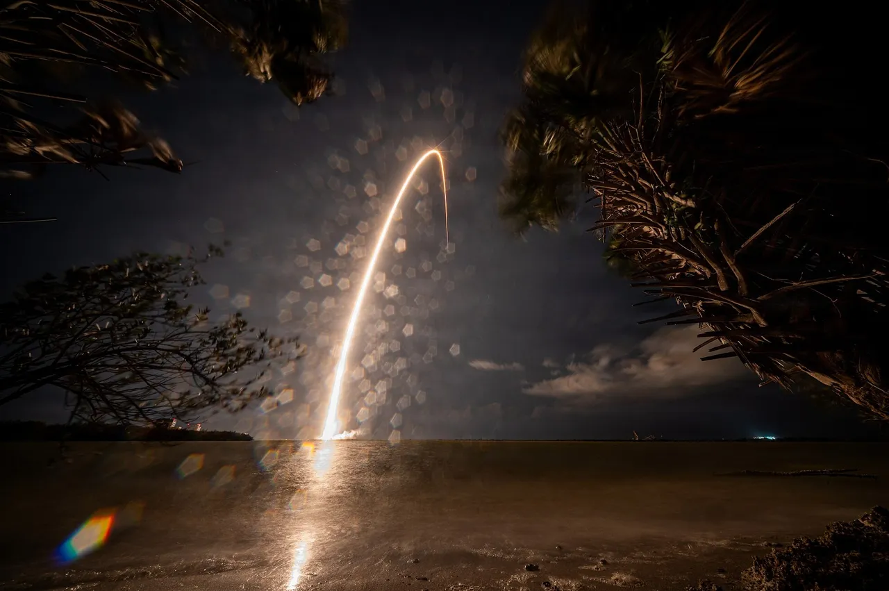 A rocket rises on a bright plume of fire and smoke from a coastal launch pad under a blue sky
