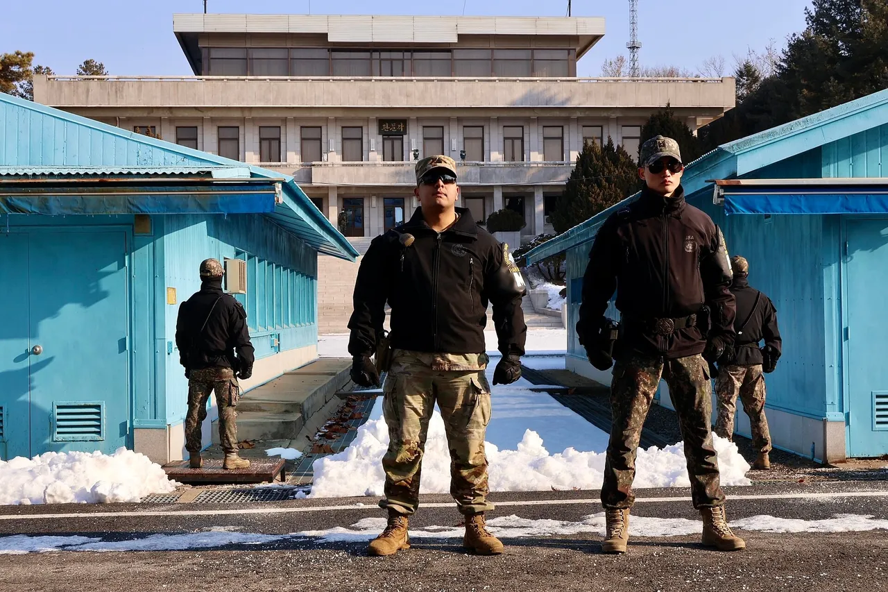 Two soldiers in camouflage trousers, dark jackets and sunglasses stand between blue huts on snowy ground, with a large grey building behind them