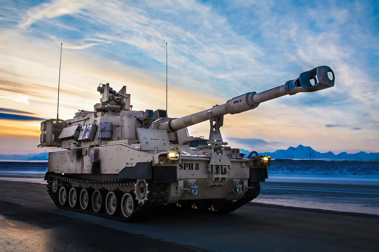 A tan tracked M109A7 self-propelled howitzer with its gun raised stands on a road at dusk, with snowy mountains behind it.