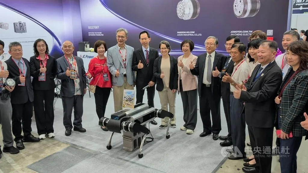 Hsiao Bi-khim and exhibitors stand behind a black Taiwan Tu Gou robot dog on a pedestal at an industrial trade show booth.