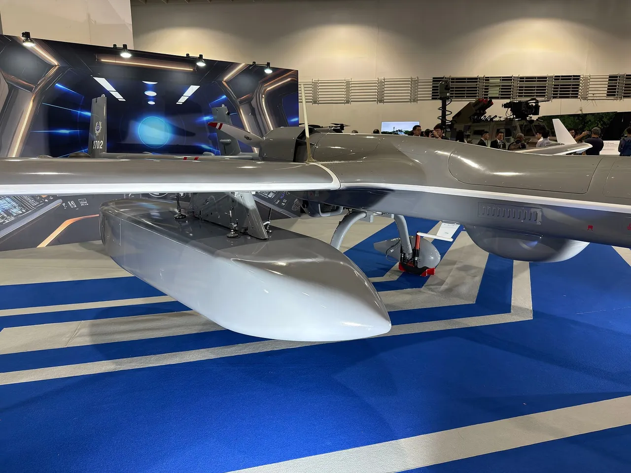 Close view of a grey military drone with a large white pod mounted under its wing on a blue exhibition floor