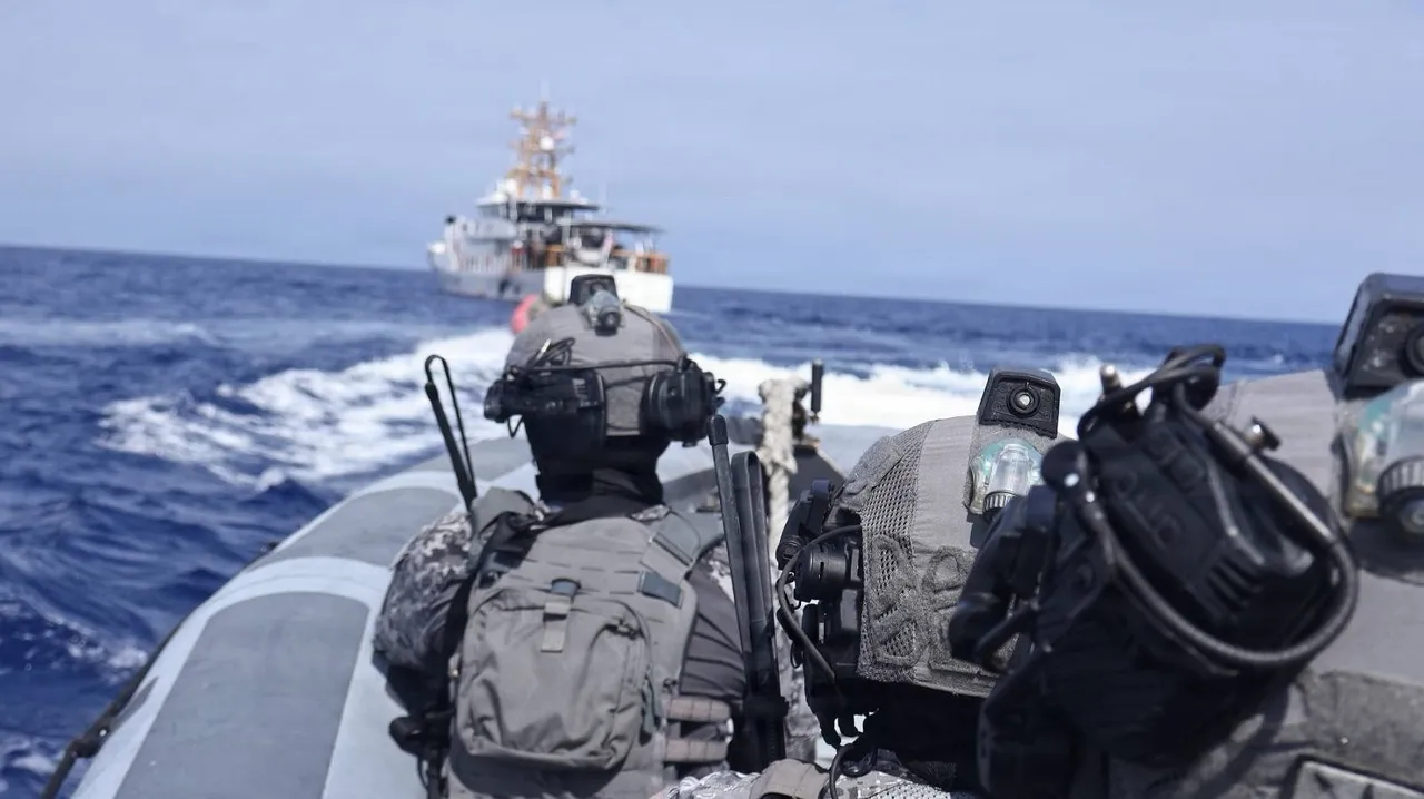 Helmeted coast guard boarding team members in body armour sit in a grey rubber boat speeding toward a white patrol cutter on open blue sea