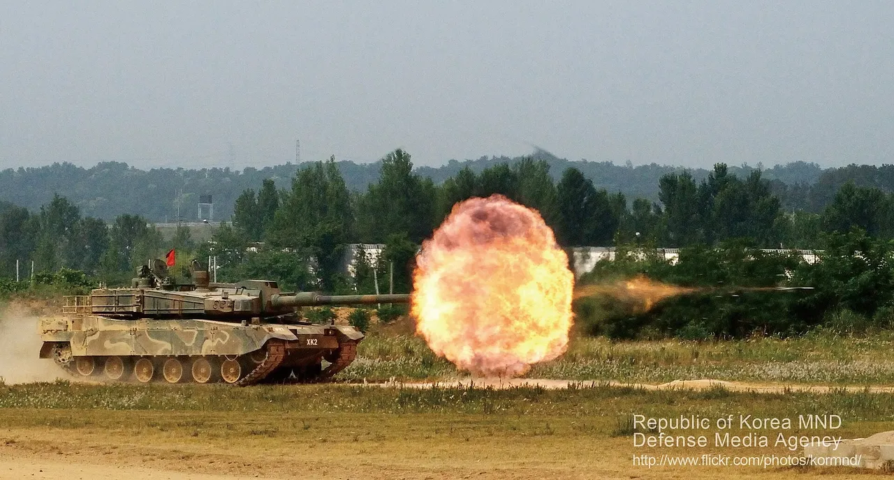 A camouflaged K2 battle tank fires its main gun, producing a large fireball across an open range