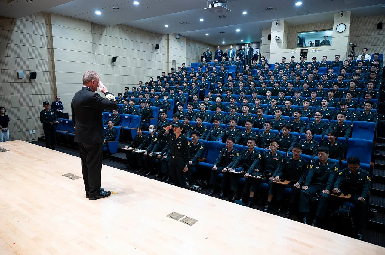 Rows of Korea Military Academy cadets in dark uniforms seated in an auditorium as a visitor salutes them from the stage