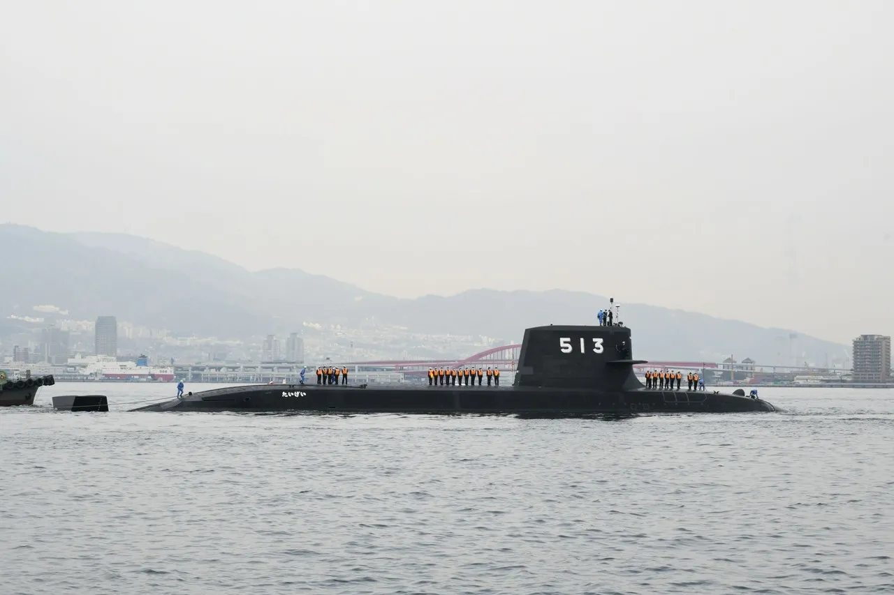 A black Japanese submarine with the hull number 513 on its sail sails through a harbour with crew lined up on deck and hills behind
