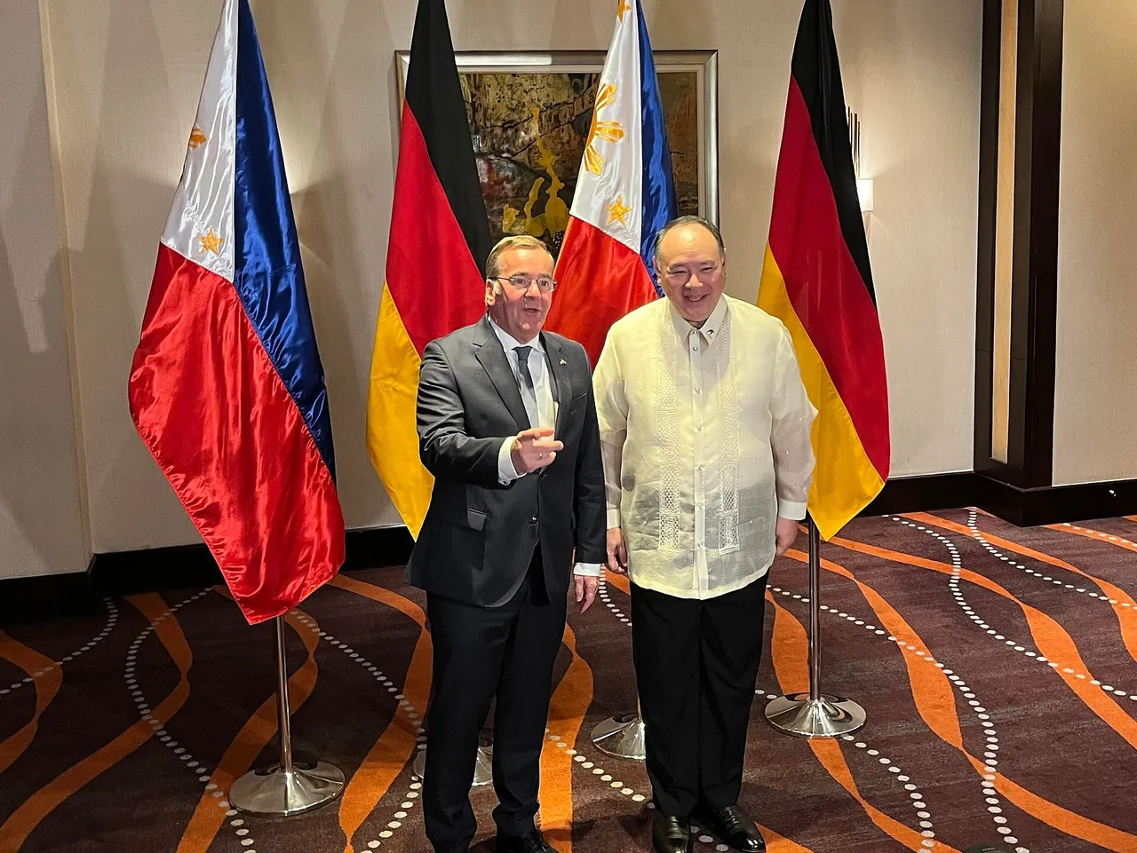 Two men in front of Philippine and German flags, one in a dark suit and one in a white barong