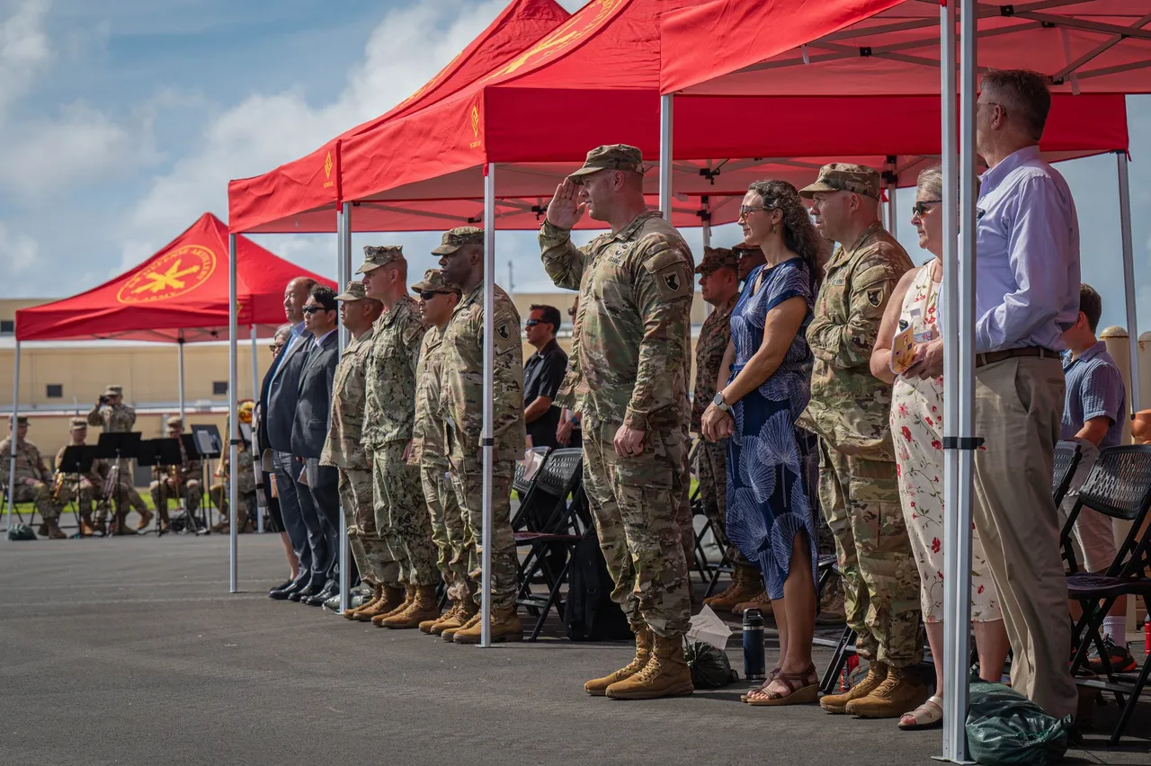 US Army soldiers in camouflage uniforms, one saluting, stand in a line with civilian guests under red tents at an outdoor ceremony