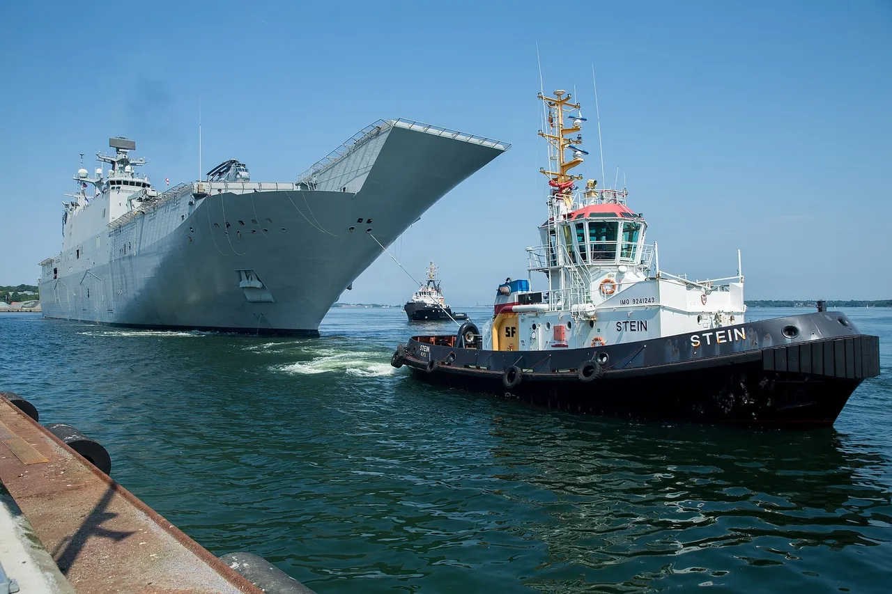 A large grey amphibious assault ship with a ski-jump flight deck is guided into harbour by a black and white tugboat under a clear blue sky.