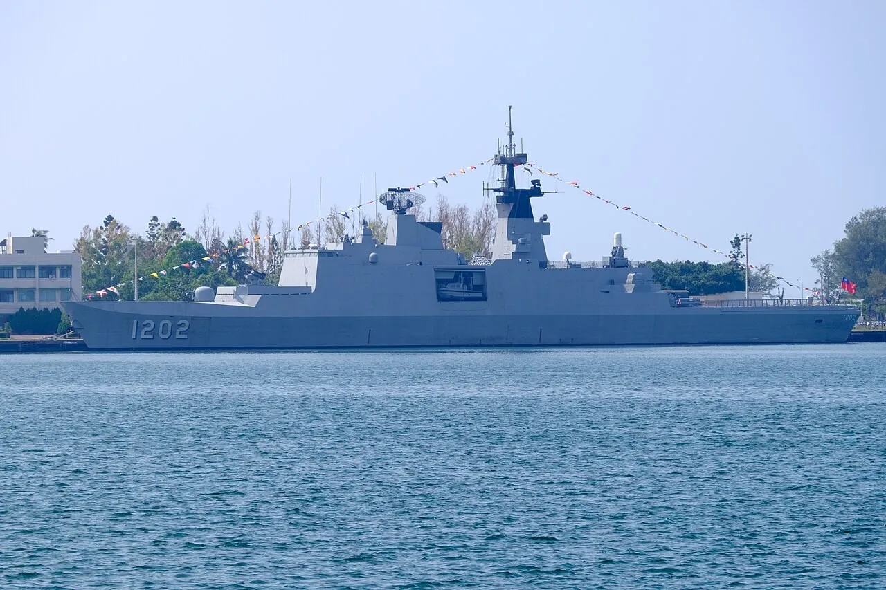 A grey angular warship with the hull number 1202 and signal flags strung from its mast, moored beside a pier with trees and buildings behind it.