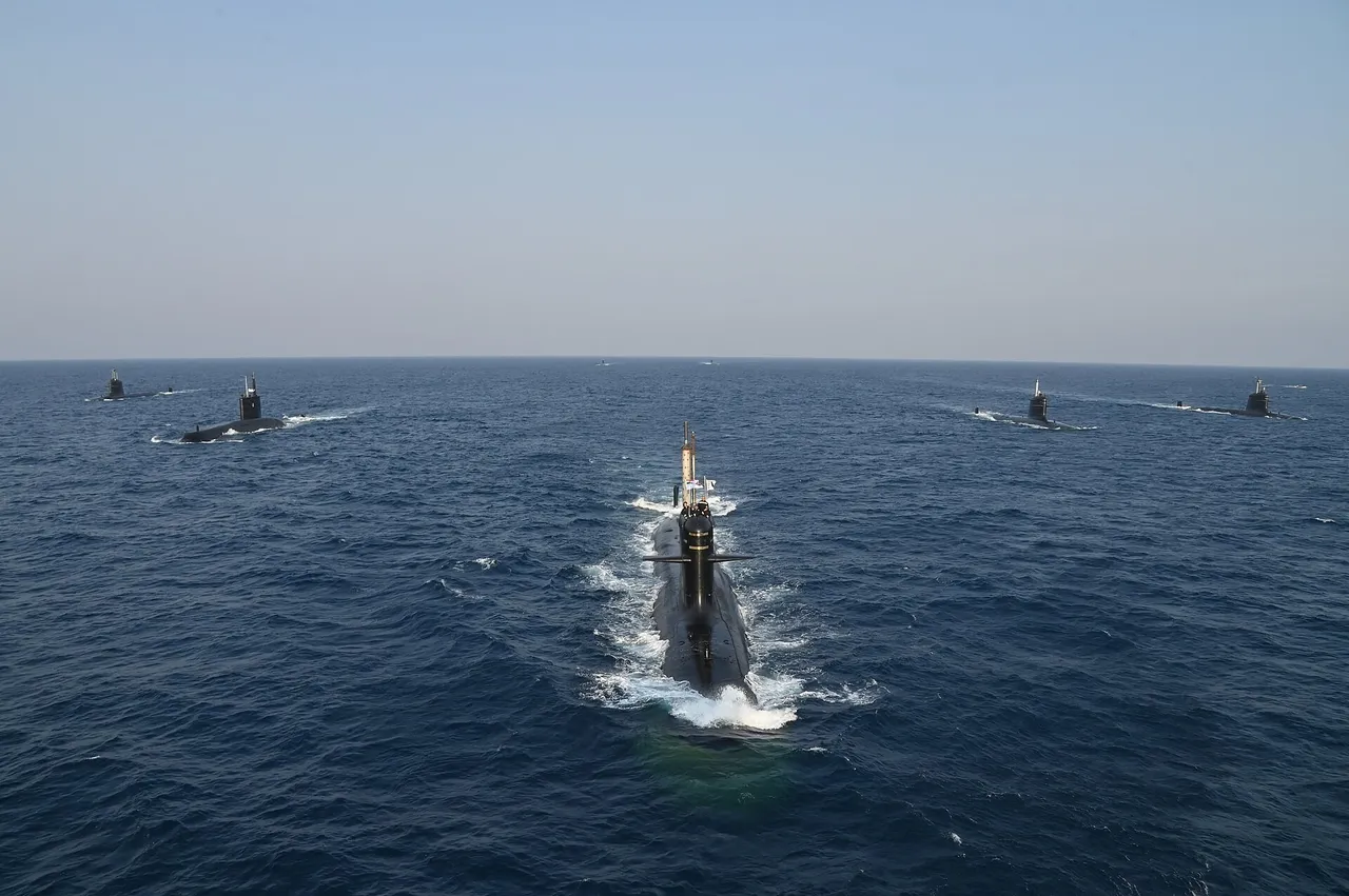 A black submarine sails on the surface toward the camera, leaving a white wake, with several other submarines spread out behind it on a calm blue sea