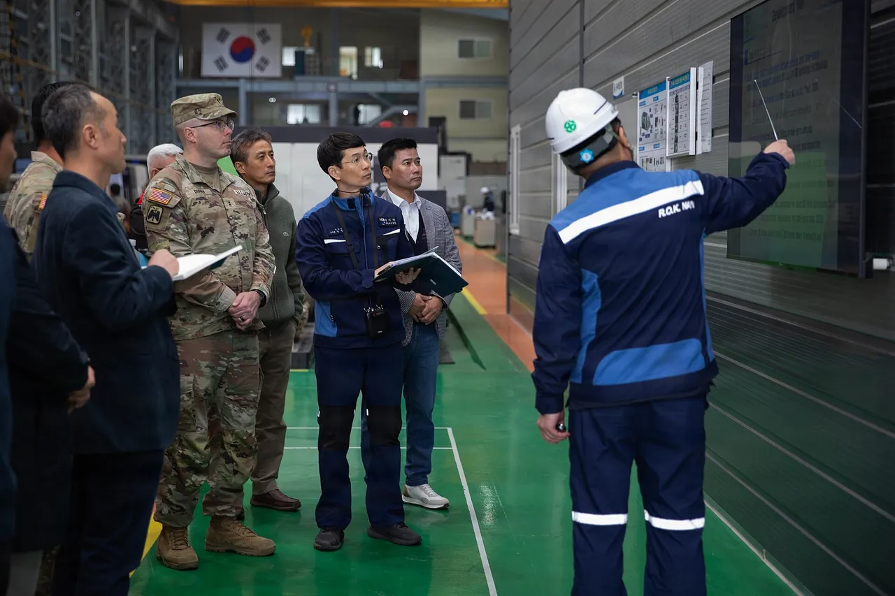 US Army officers in uniform and civilian clothes listen as a South Korean Navy worker in a hard hat points to a display inside a factory hall.