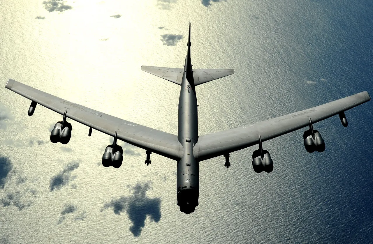 A large grey eight-engine bomber with swept wings seen from above, flying over a sunlit ocean with scattered clouds below