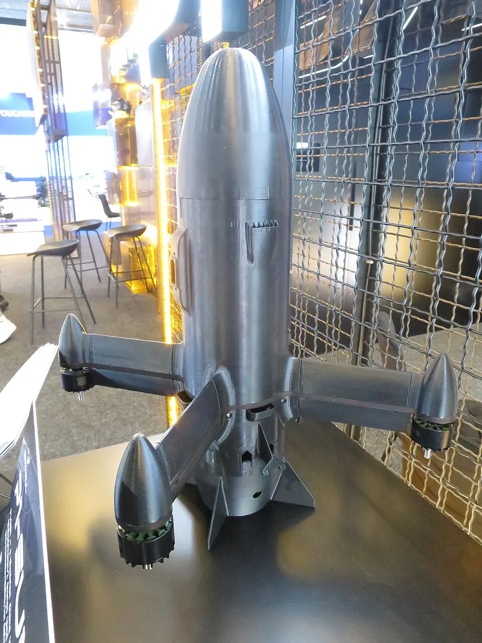 A dark grey, bullet-shaped interceptor drone with four short arms ending in propeller motors, displayed nose-up on a stand in an exhibition hall