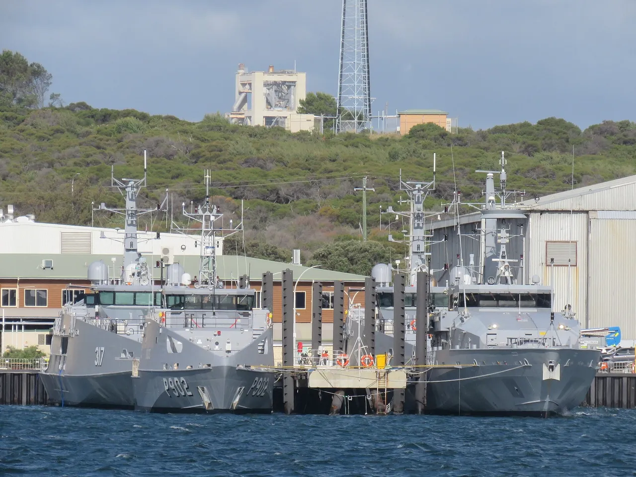 Several grey and white patrol boats moored side by side at a shipyard quay with large sheds behind them