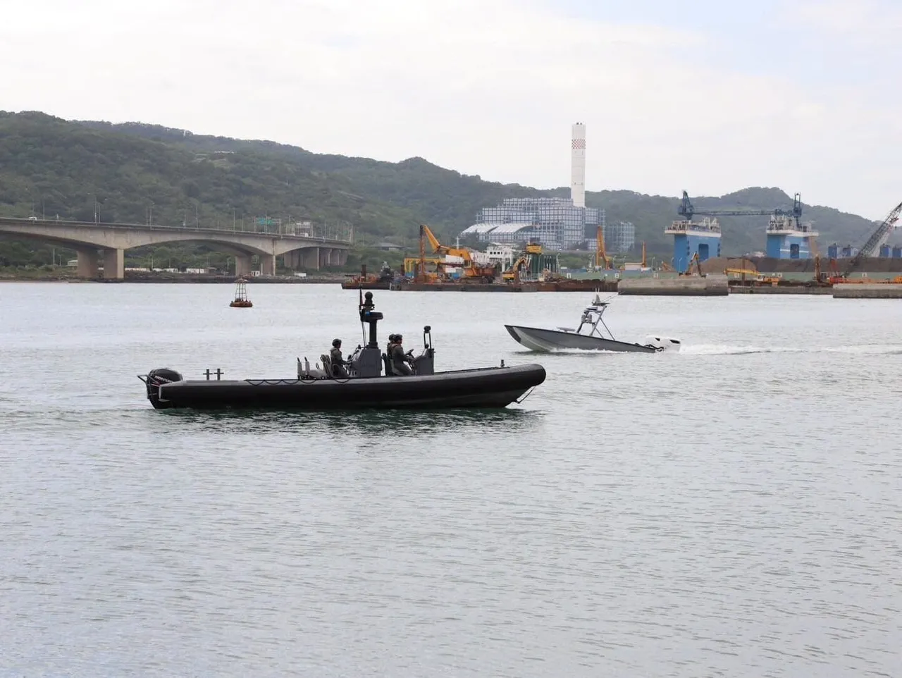 A grey unmanned surface vessel moves across a harbour near a black rigid-hull boat carrying coast guard personnel, with cranes and hills behind.