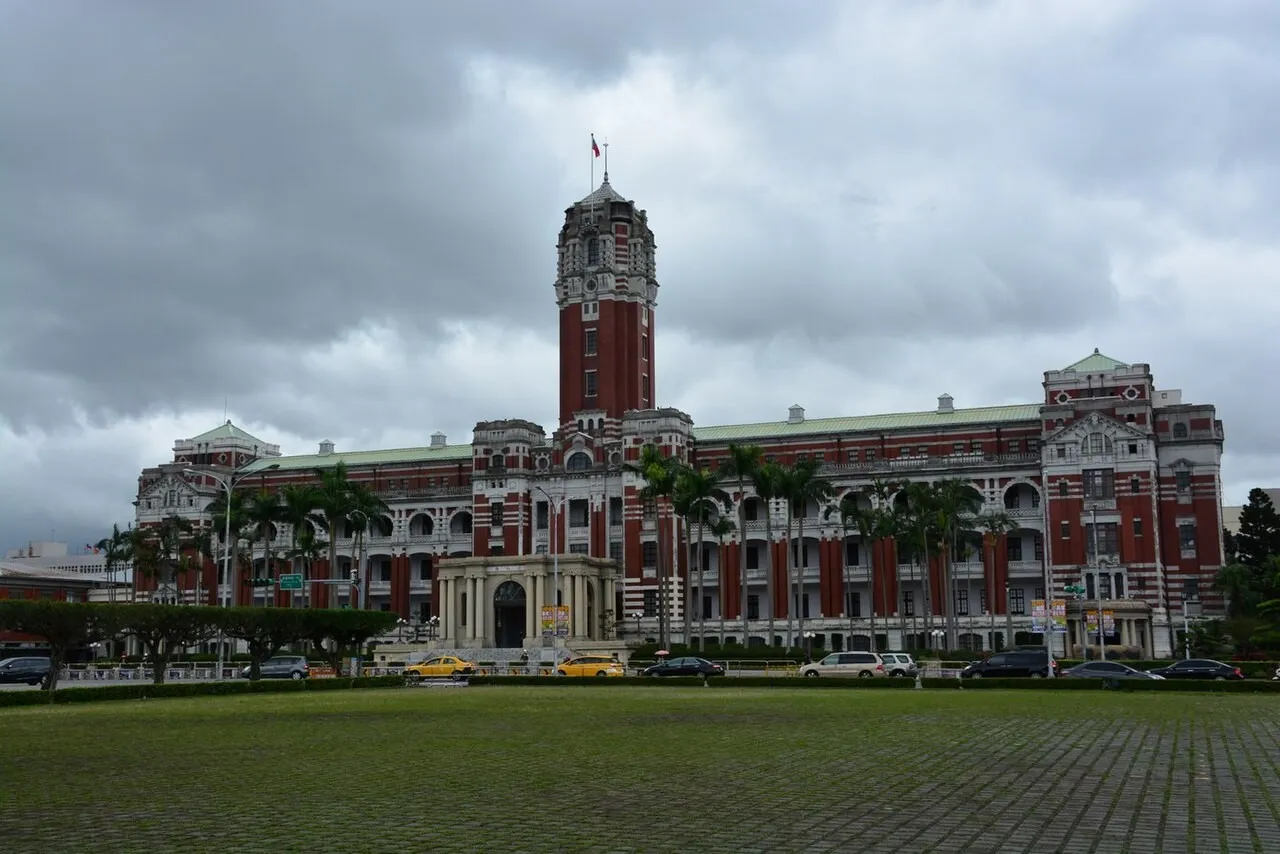A red-brick and white-stone colonial-era building with a tall central clock tower flying a flag, palm trees along its front and a wide lawn under a grey cloudy sky.