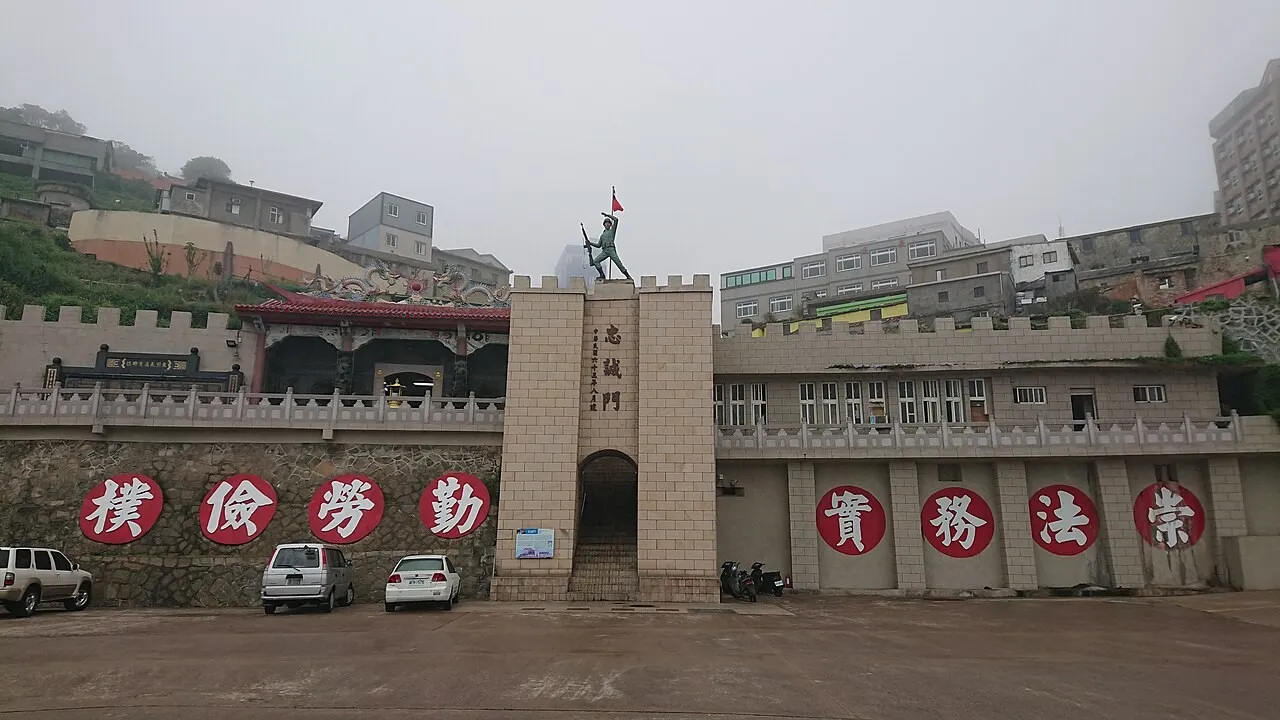 A stone gateway with crenellated walls topped by a statue of a soldier holding a flag, flanked by walls bearing large white characters on red discs, with cars parked in front and hillside houses behind under grey, misty skies.