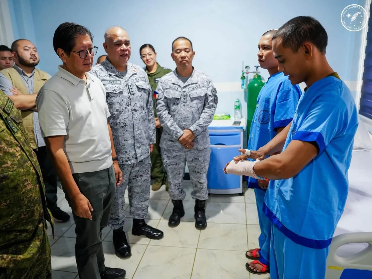 President Marcos, in a white shirt, stands beside navy officers as two injured sailors in blue hospital gowns show a bandaged hand.