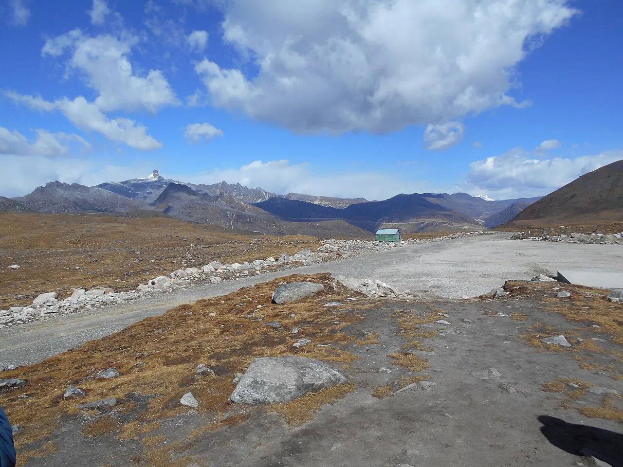 A gravel road crossing a high, barren mountain pass under a blue sky with snow-capped peaks behind and a small green-roofed hut in the distance