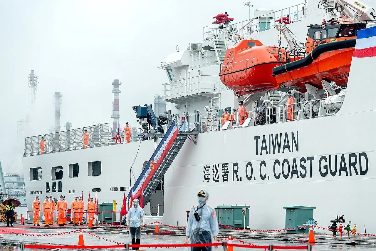 A large white ship marked Taiwan R.O.C. Coast Guard with orange lifeboats, crew in orange uniforms standing along its rails and on the quay under a grey sky.
