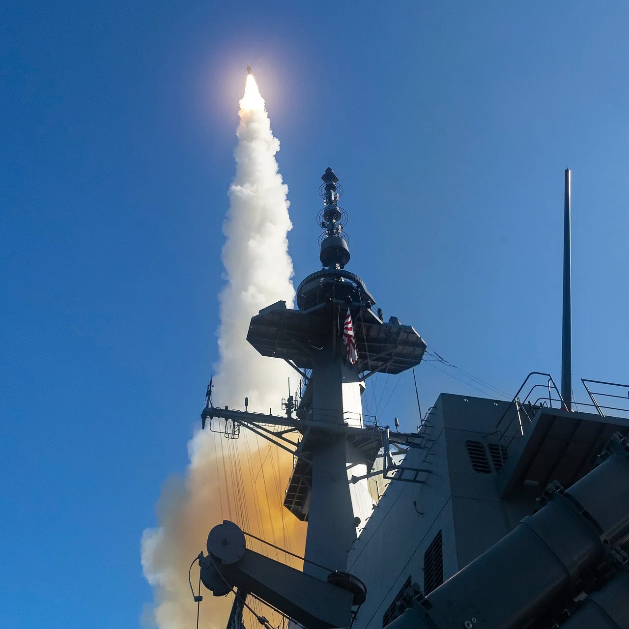 A missile trailing a white and orange exhaust plume climbs into a clear blue sky beside the grey mast of a warship