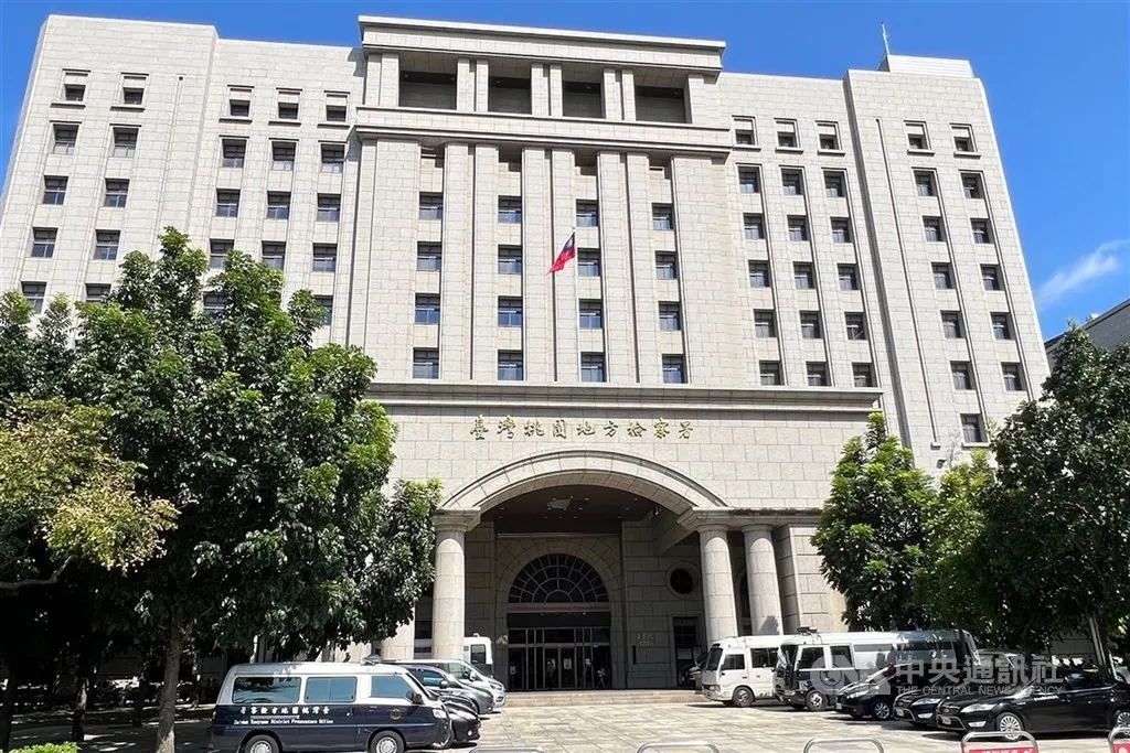 Exterior of a tall pale stone government building with a Taiwanese flag above an arched entrance, trees in front and vans parked in the forecourt.