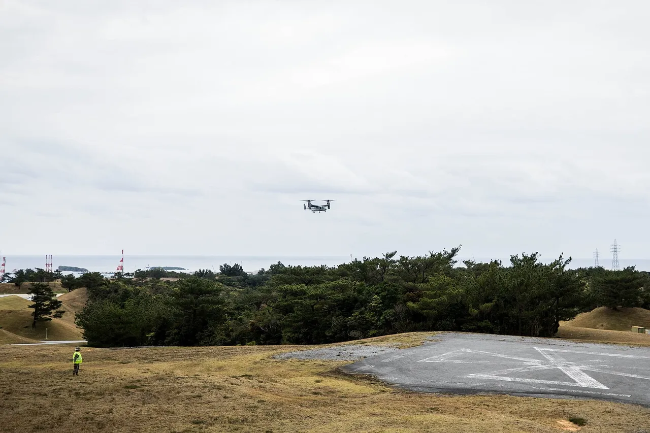 A tiltrotor aircraft flies low over pine trees towards a grey concrete helicopter pad on a grassy hill, with the sea on the horizon