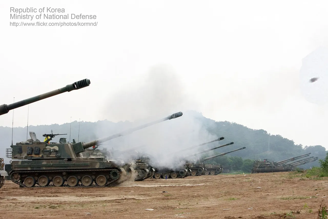 A line of tracked K9 self-propelled howitzers fire their long barrels on a dusty field, with gun smoke drifting towards wooded hills