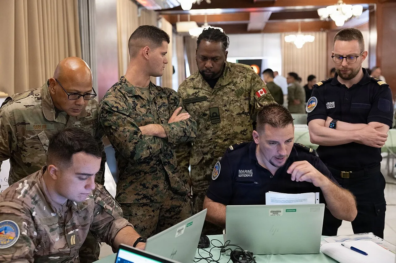Service members in French Navy uniforms and camouflage fatigues gather around laptops on a table, watching a French sailor work at a computer.