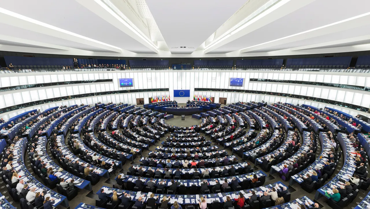 Wide view of the European Parliament's semicircular plenary chamber in Strasbourg, with members seated in curved rows facing the EU flag.