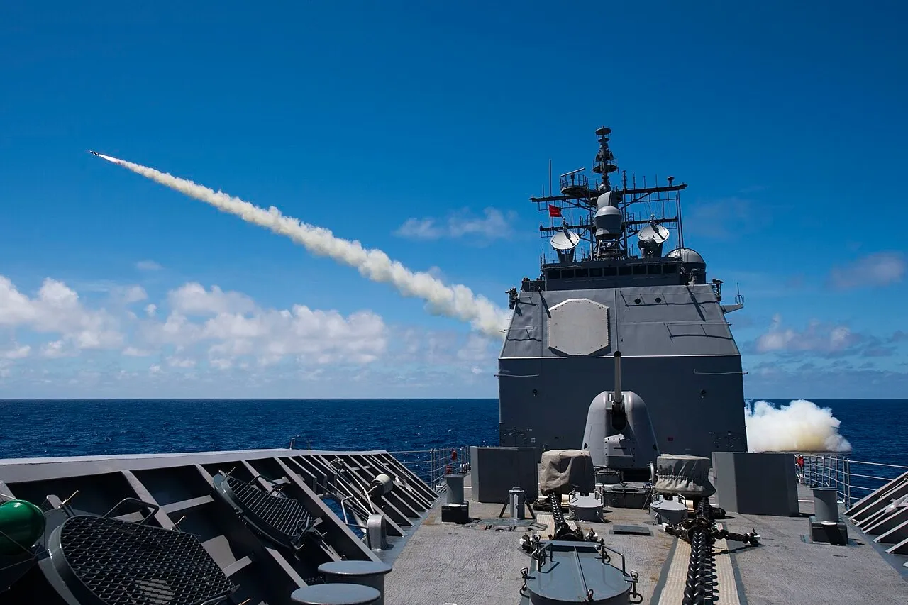 A missile trails a white smoke plume into a blue sky after launching from a grey warship, seen from the ship's foredeck with a deck gun in the foreground and open sea behind.