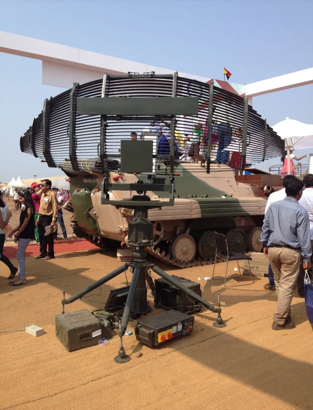 A tripod-mounted Bharani low-level radar with a curved grille antenna displayed outdoors in front of an armoured vehicle as visitors walk past
