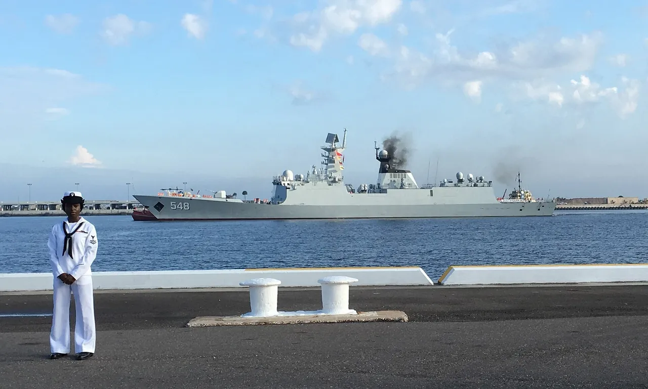 A grey Chinese navy frigate with pennant number 548 sails into a harbour while a US Navy sailor in white uniform stands on the pier in the foreground