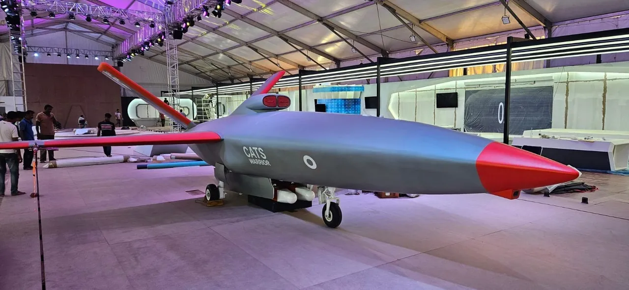 A grey drone model with a red nose and red-tipped wings sits on its landing gear inside a large white exhibition hall