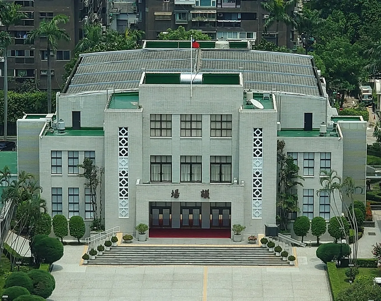 A pale grey multi-storey legislative hall with a flagpole on the roof and a wide staircase leading to its entrance, surrounded by trees and apartment blocks.
