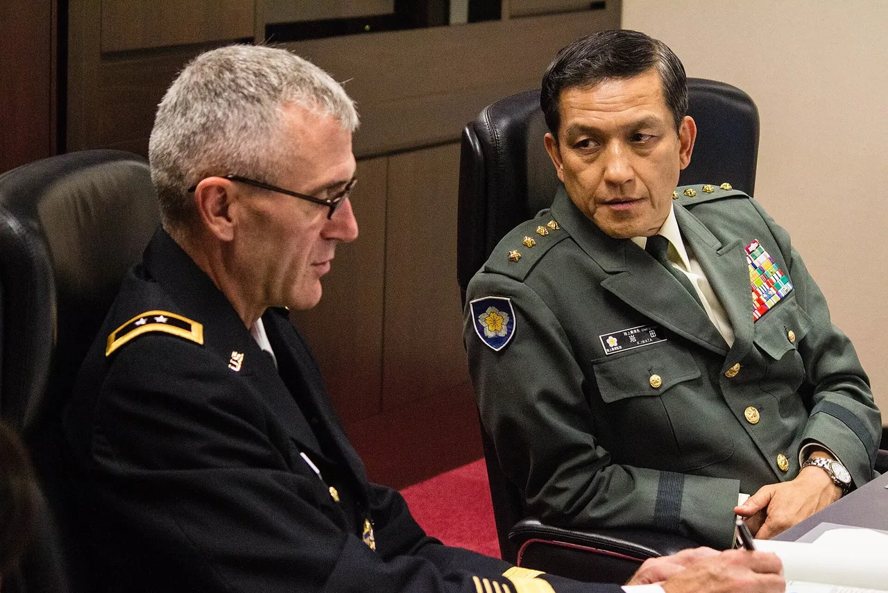 A Japanese general in a green uniform sits at a conference table beside a US Army general in a dark uniform.