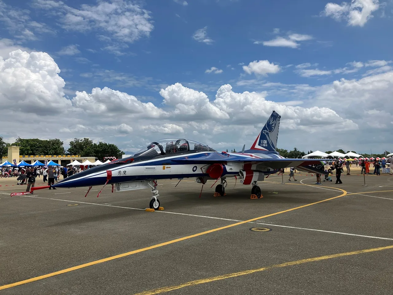 A blue, white and red Brave Eagle jet trainer parked on an air base apron with red engine intake covers fitted