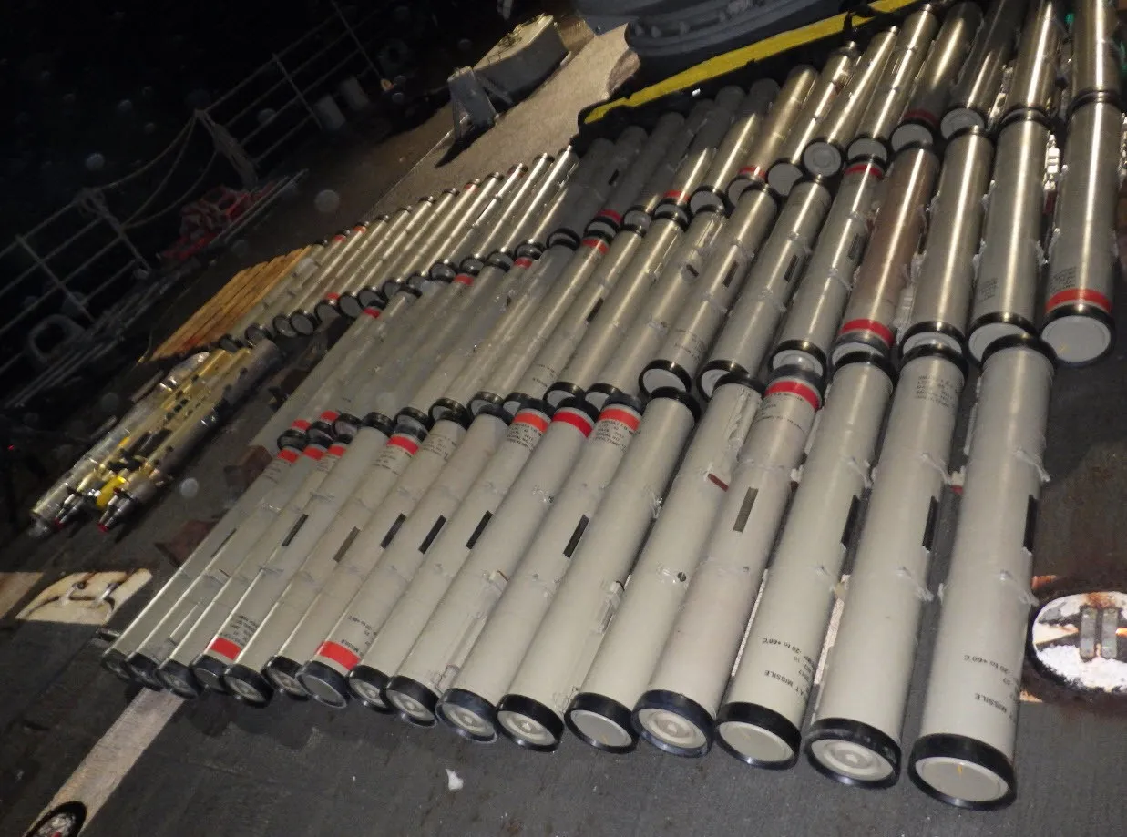 Rows of pale missile tubes with red bands laid out on a warship deck at night beside other weapon parts