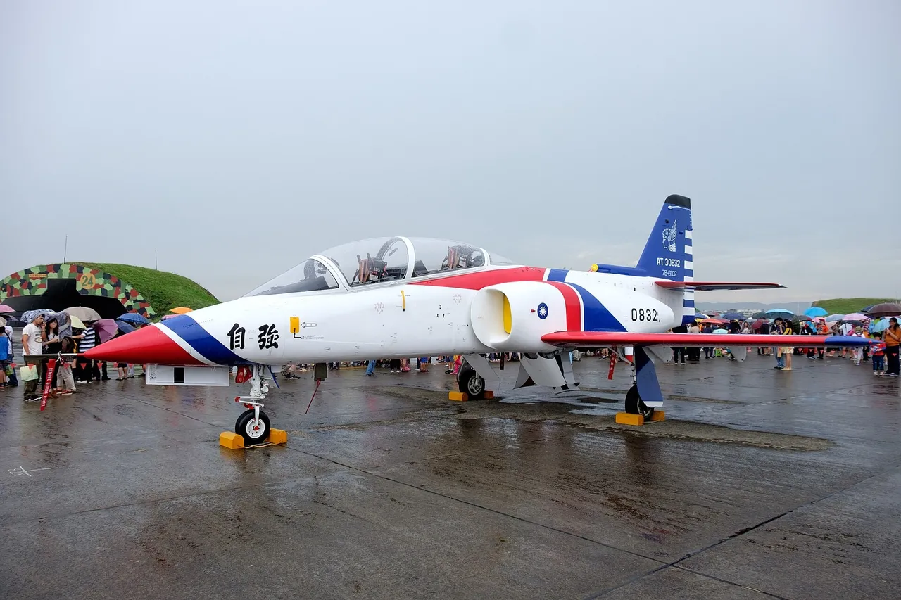A white two-seat jet trainer with red and blue stripes parked on a wet air base apron as visitors with umbrellas look on