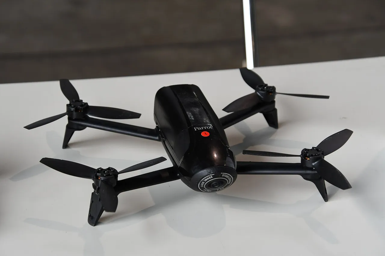 A small black four-rotor Parrot quadcopter drone with a front camera lens resting on a white table