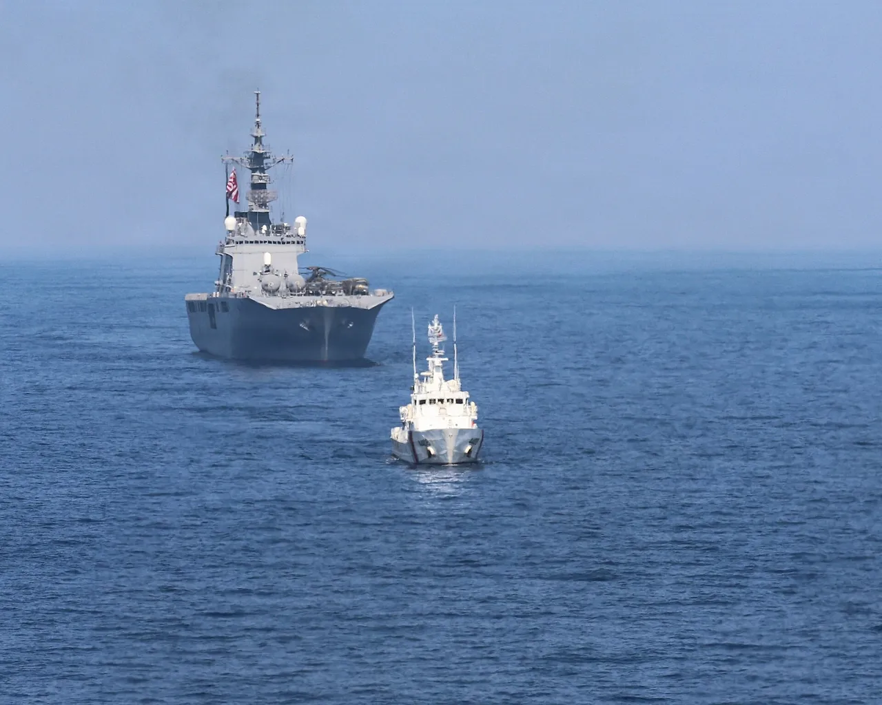 A small white Philippine Coast Guard patrol boat sails in front of a large grey Japanese navy landing ship on open sea
