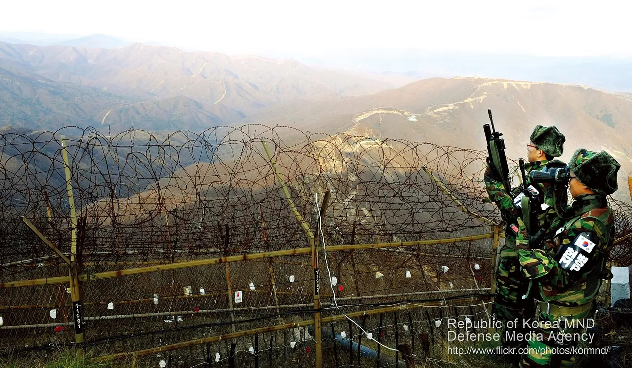 Two South Korean soldiers in winter camouflage with rifles and binoculars stand beside a barbed-wire fence overlooking mountain ridges